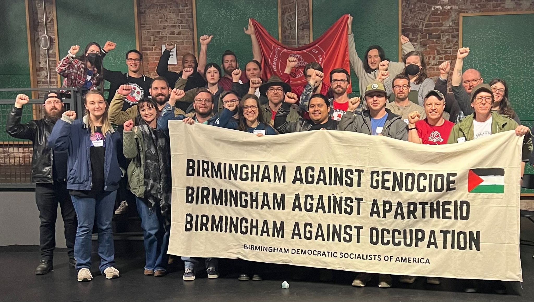 DSA and YDSA members from Alabama and Mississippi pose with fists raised behind a banner with a Palestinian flag that reads 'Birmingham Against Genocide, Birmingham Against Apartheid, Birmingham Against Occupation, Birmingham Democratic Socialists of America.' Behind them is a red DSA flag.